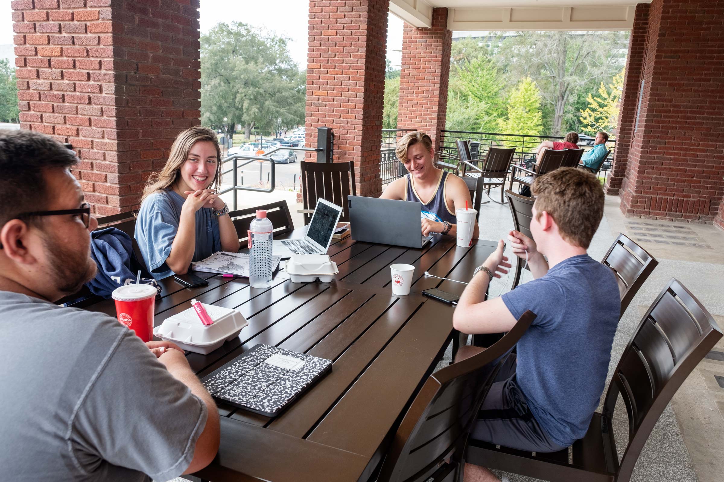 Four students chat and eat lunch between classes at a table on the YMCA's porch.