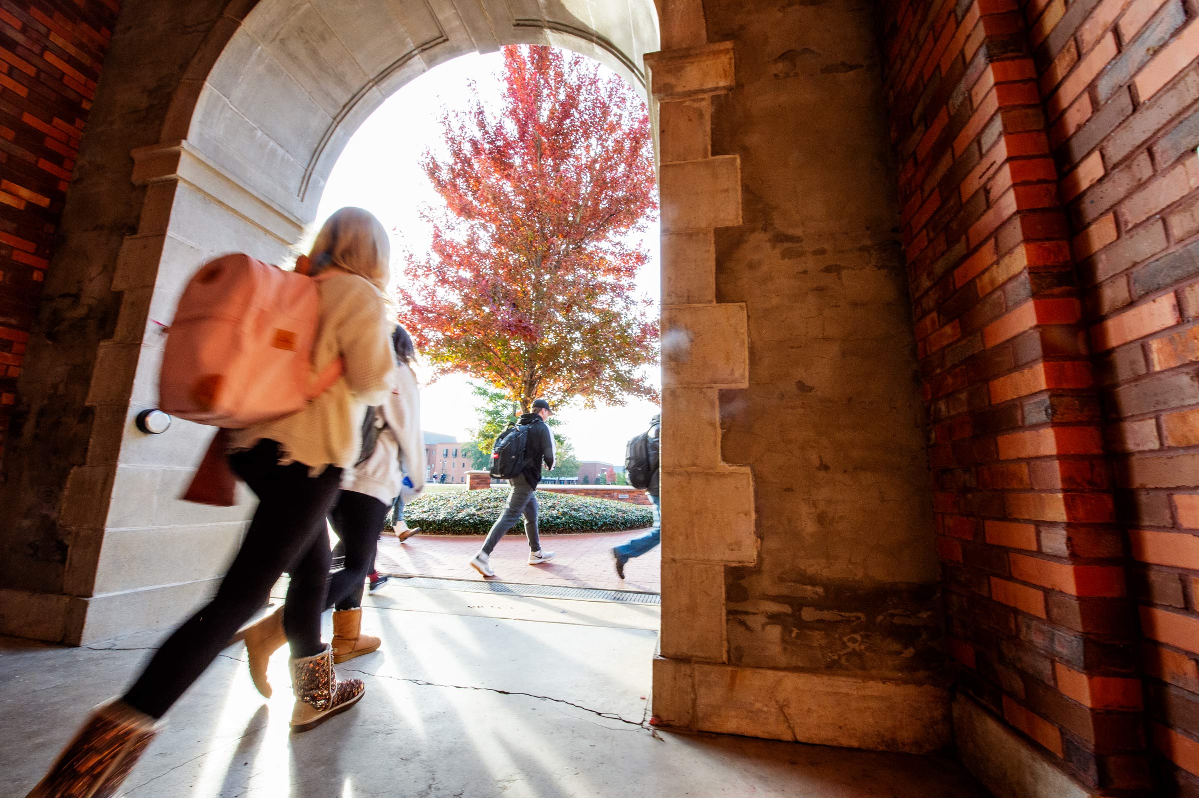 Students walk out of the McCain Hall arch with a maple tree's fall leaves framed in the doorway.