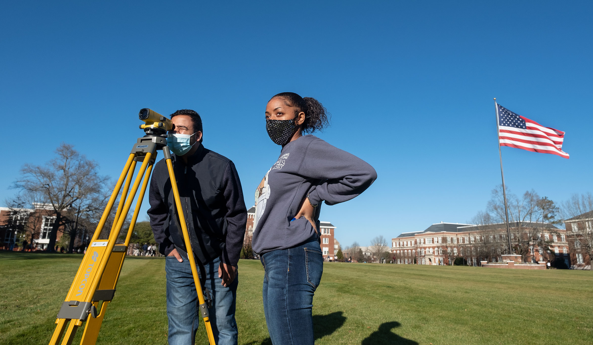 With a sunlit Drill Field in the background, Engineering student Nim Kc leans in to look through the survey viewfinder while Courtlyn Carson looks on. Both wear masks for safety against Covid-19.