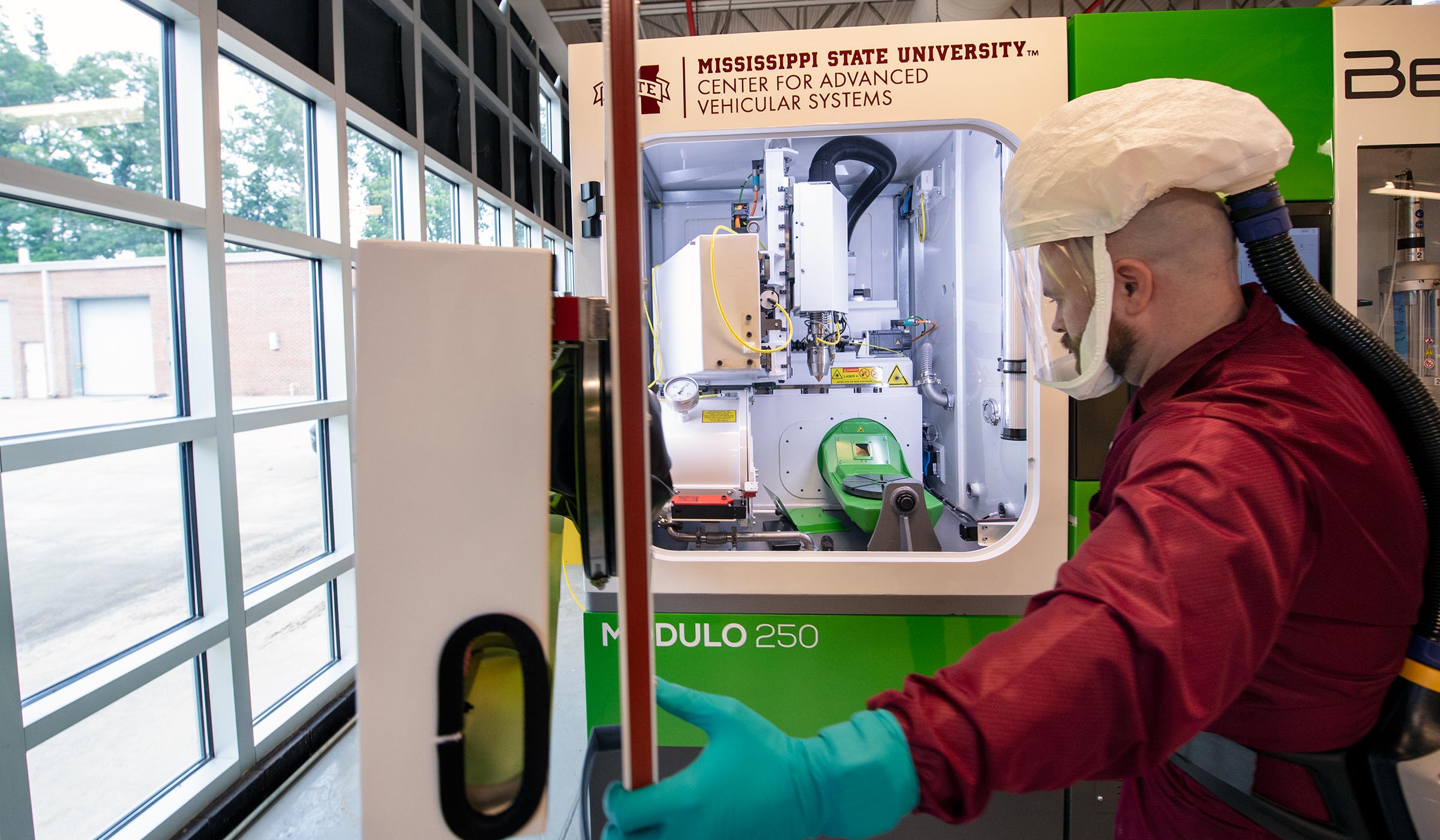 CAVS Research Engineer - wearing full face clear protective lab face covering - opens door to Additive Manufacturing Lab's large BeAM Modulo 250 machine, which is white and green.