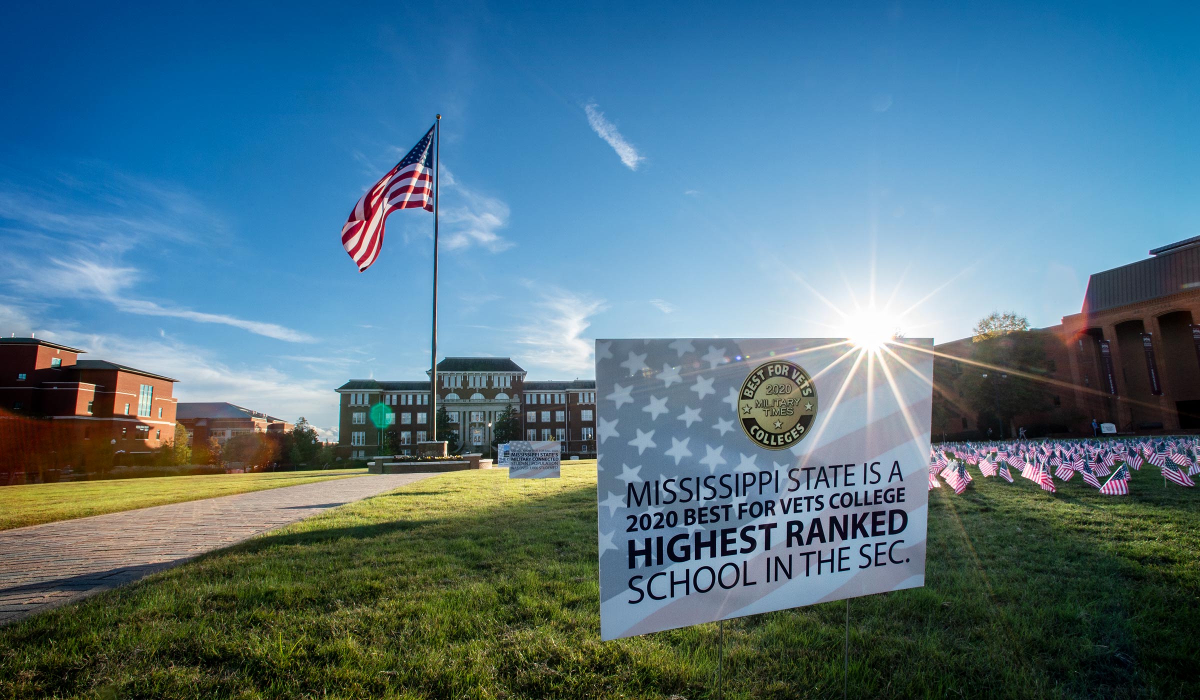 Sunflare backlights sign on Drill Field next to Veterans Day American flags: MSU is a "2020 Best for Vets College Highest Ranked School in the SEC."