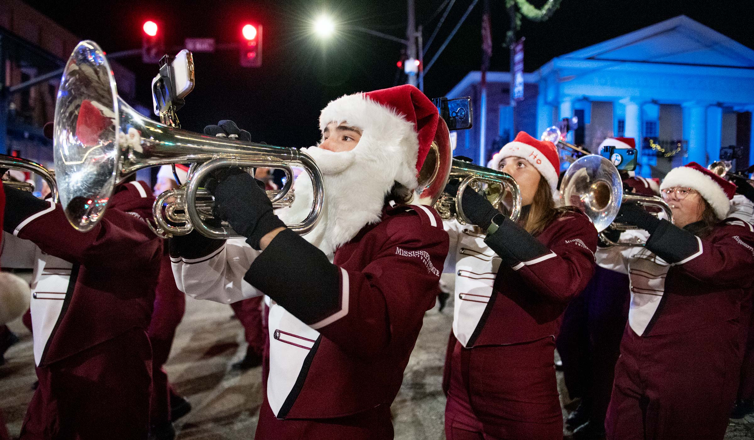Wearing a Santa hat and white beard, a band member marches past Starkville downtown's courthouse at night with his fellow mellophone players.