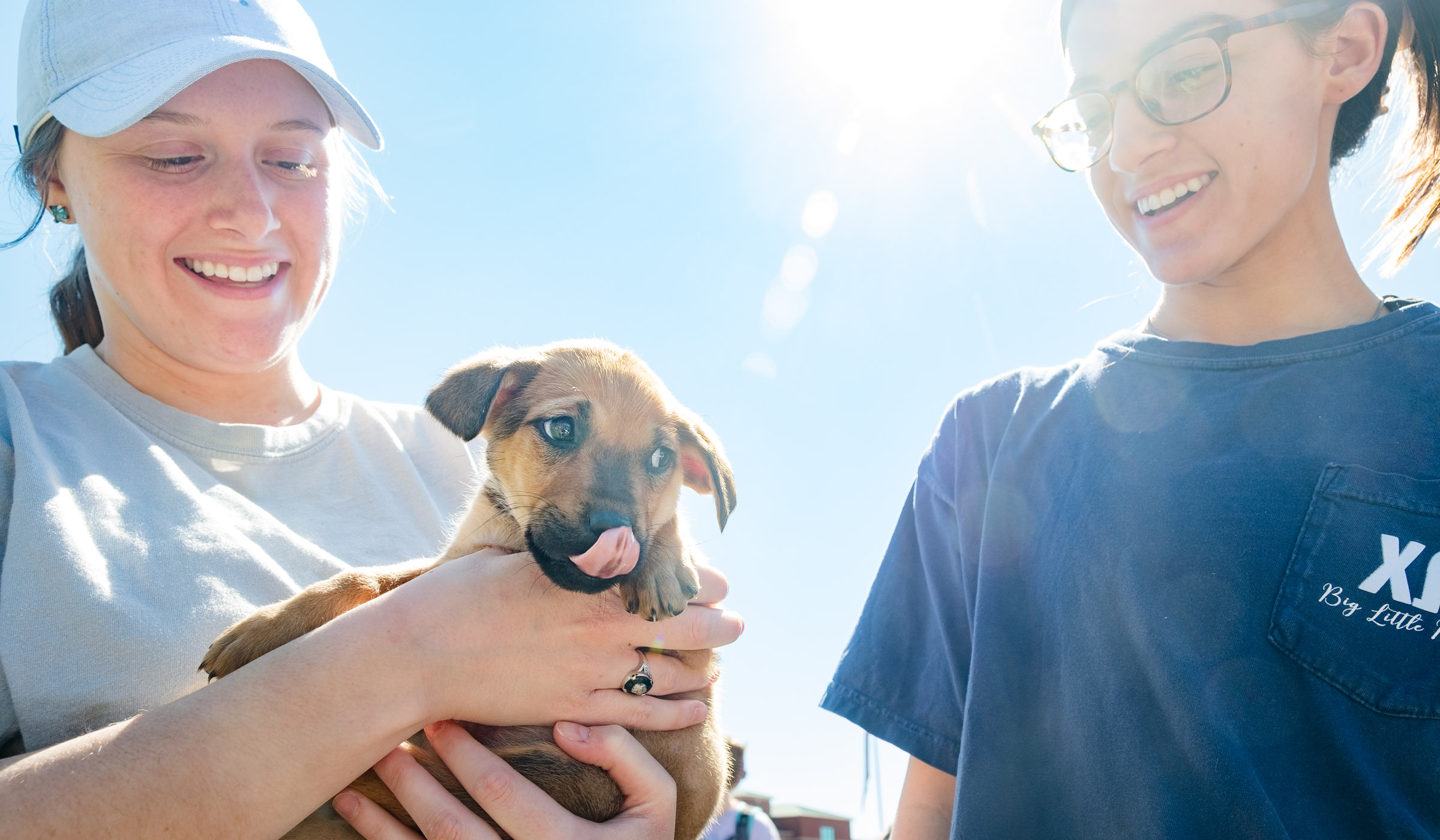 With a small puppy held in the center of the frame, two female students look on smiling with blue sky behind them.