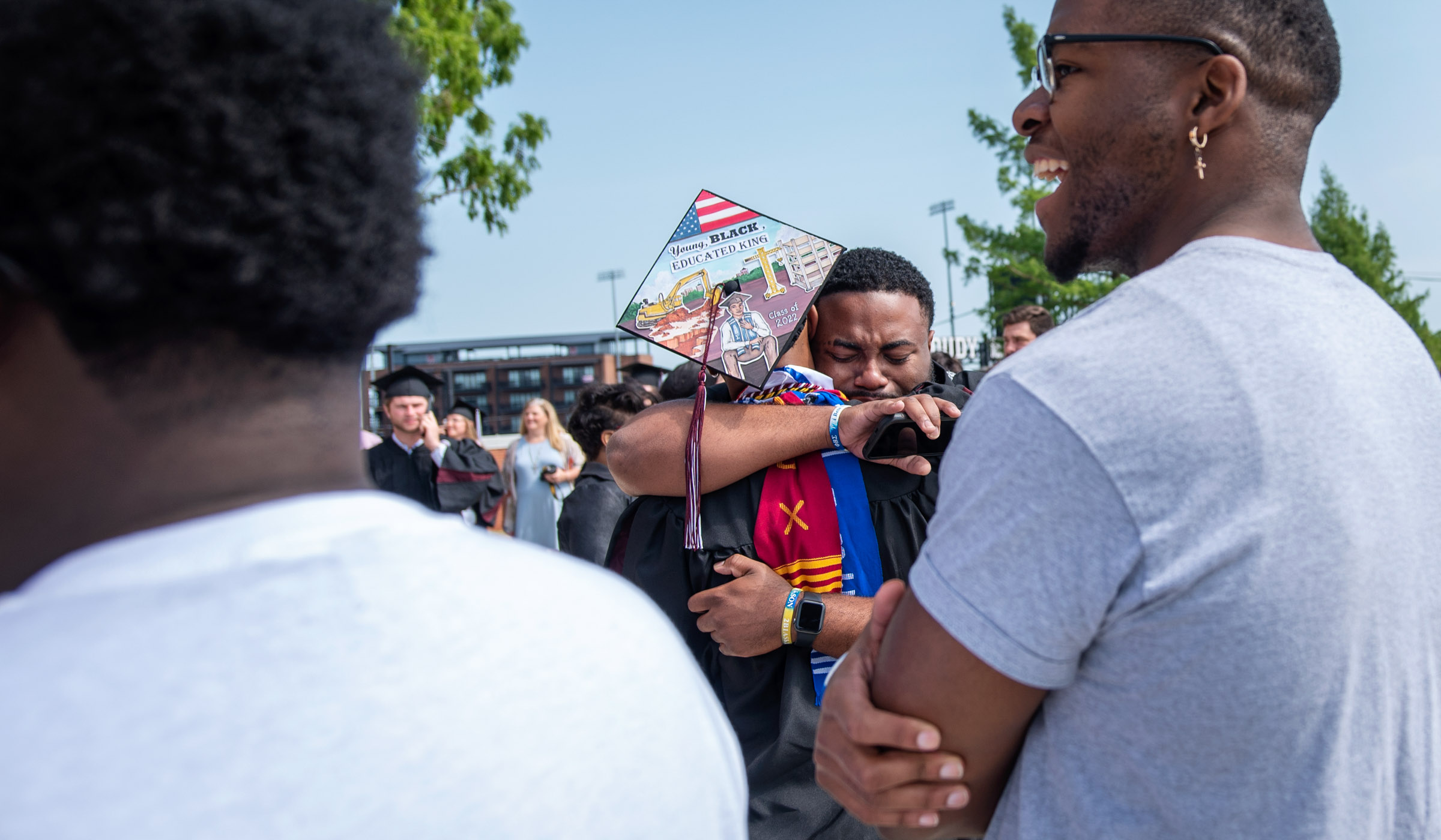 Framed by the shoulders of fellow graduation guests, one student hugs a graduate whose back is turned to the camera, displaying the decorated mortar board he's wearing.