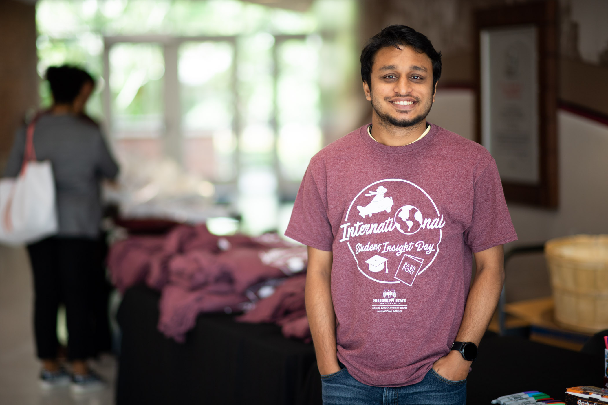 An incoming male South Asian student wearing a light maroon "International Student Insight Day" tee smiles with hands in pockets