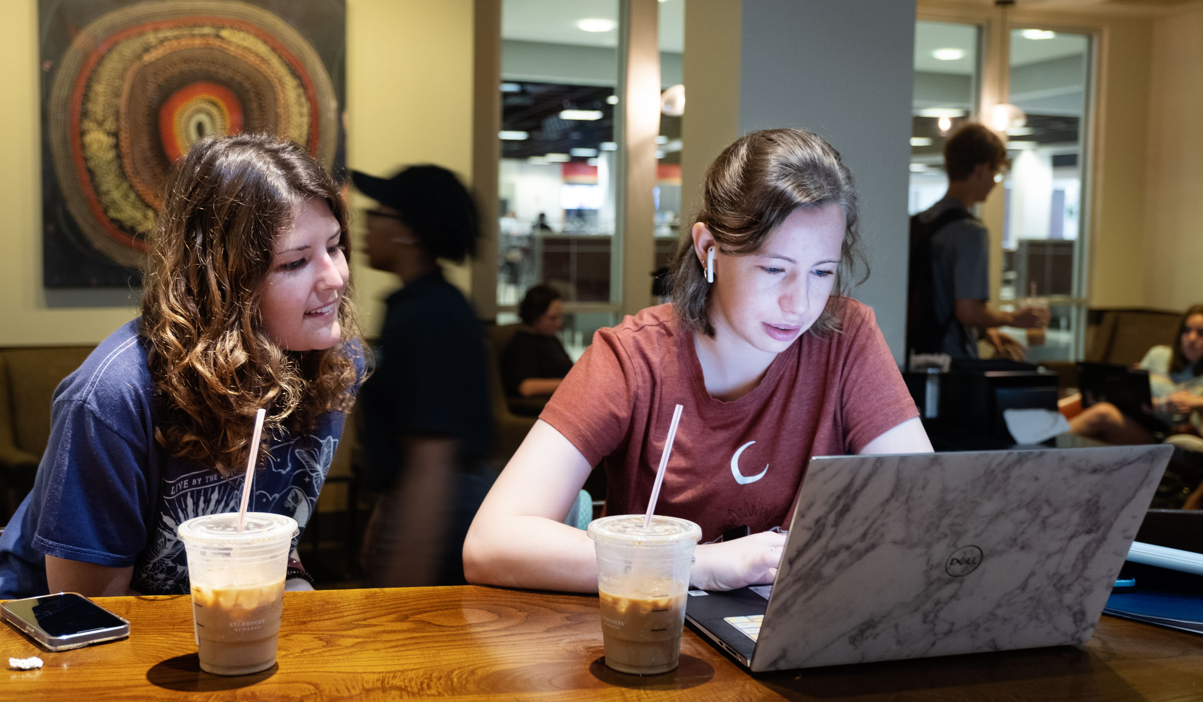 Two female students sit at the Starbucks wooden table with iced beverage, with the student on the right looking at her laptop and the other looking on. Starbucks is bustling behind them.