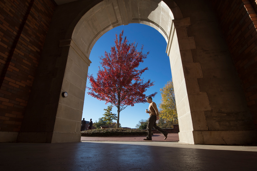 McCain archway entrance frames trees with fall leaves, blue sky, and pedestrian staff John Brocato walking past