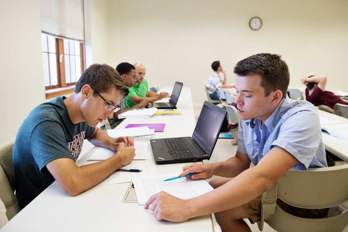 Two students discuss evaluate each other's English composition drafts at the edge of a classroom.