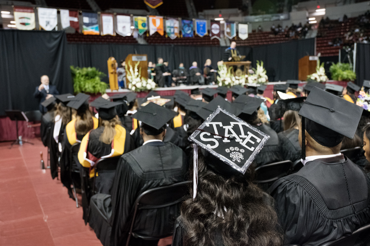 With the graduation stage in the background, a graduating student's mortar board is decorated with "State" and a pawprint.