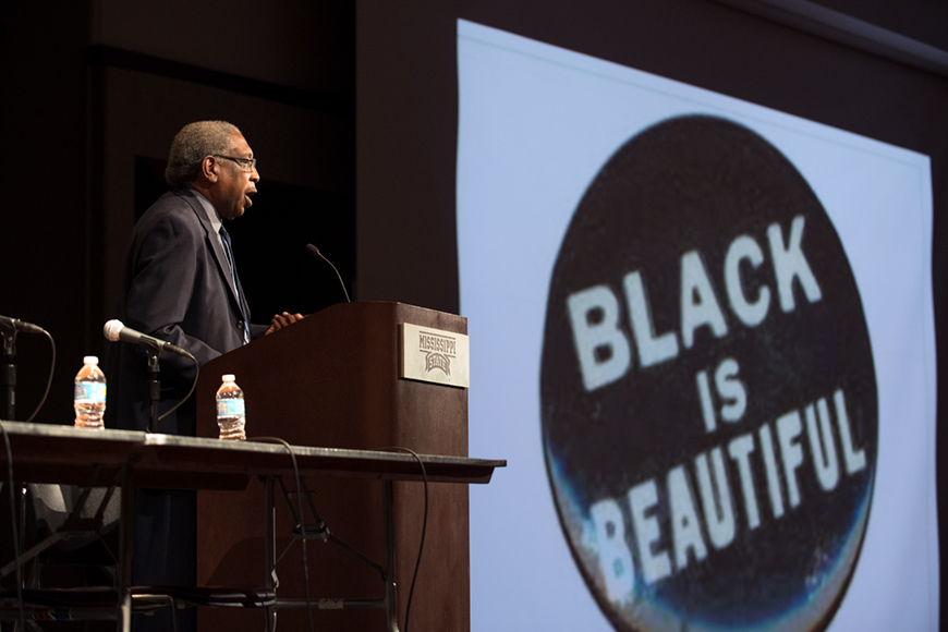 Speaker at podium with slide of "Black is Beautiful" in the background