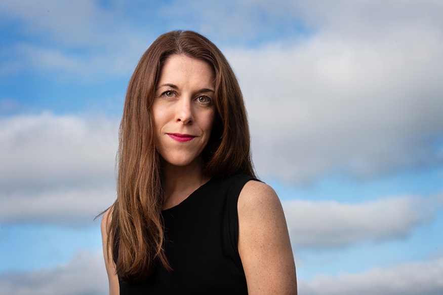 Catherine Pierce is wearing a black shirt with the blue sky and clouds behind her.